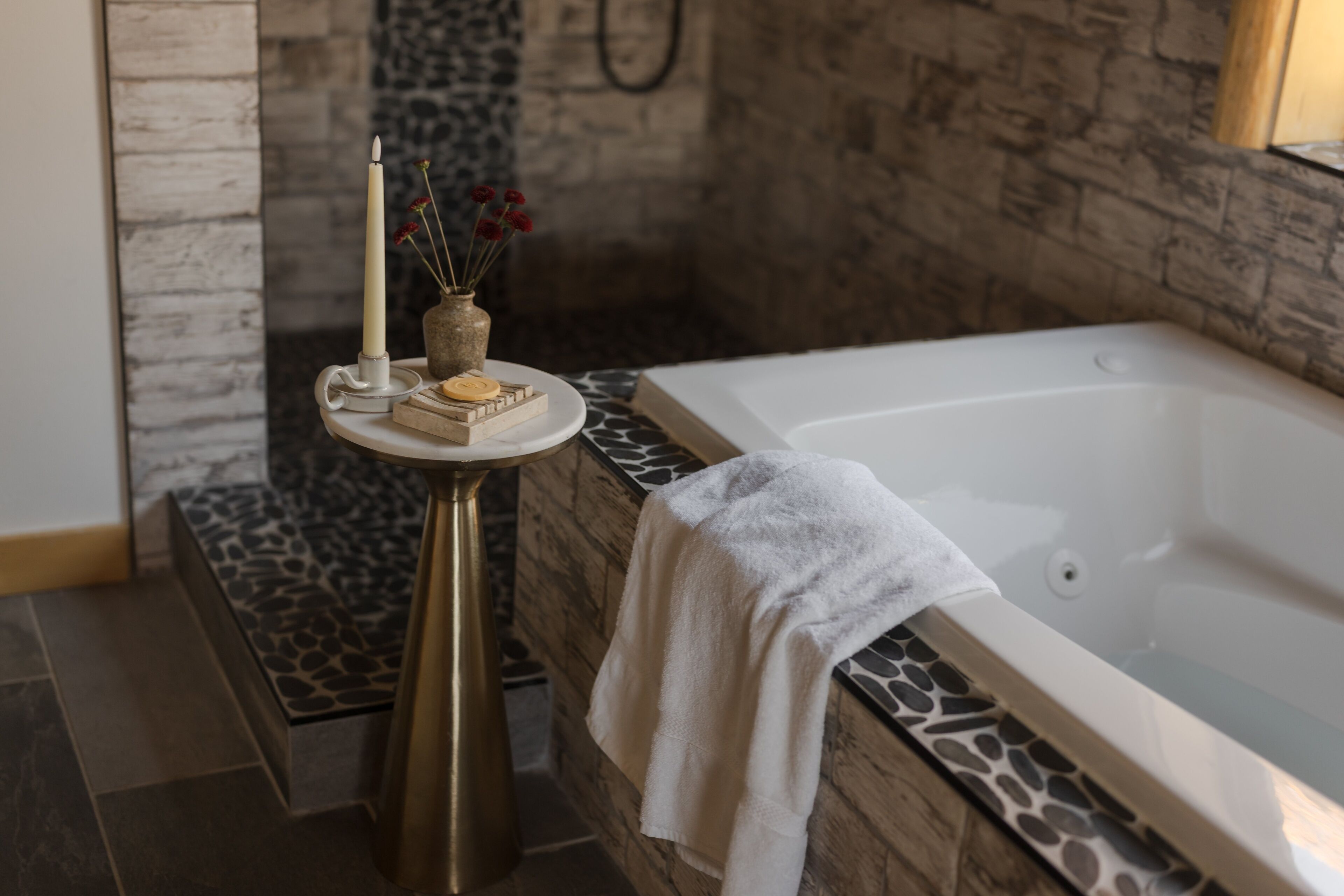 Een moderne badkamer met een stenen muur, een wit ligbad, een klein rond tafeltje met een kaars en een vaas met bloemen.