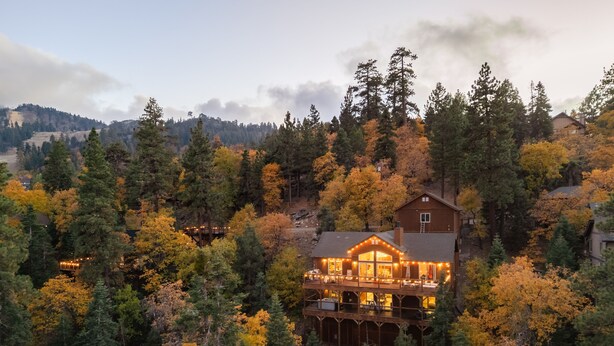 Eine Lodge mit großer Veranda und zahlreichen Fenstern, umgeben von dichtem Wald und Herbstlaub.