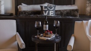 Ein gemütliches Zimmer mit einem Bett, das ein Kopfkissen mit der Aufschrift „LOVE“ und einem Herzdesign zeigt, einem kleinen Tisch mit zwei Weingläsern, einer Flasche und Erdbeeren.