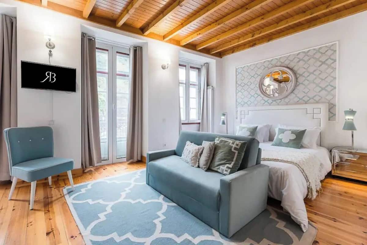 A bedroom with a blue sofa, a bed with white bedding, a TV mounted on the wall, and a patterned wall decoration.