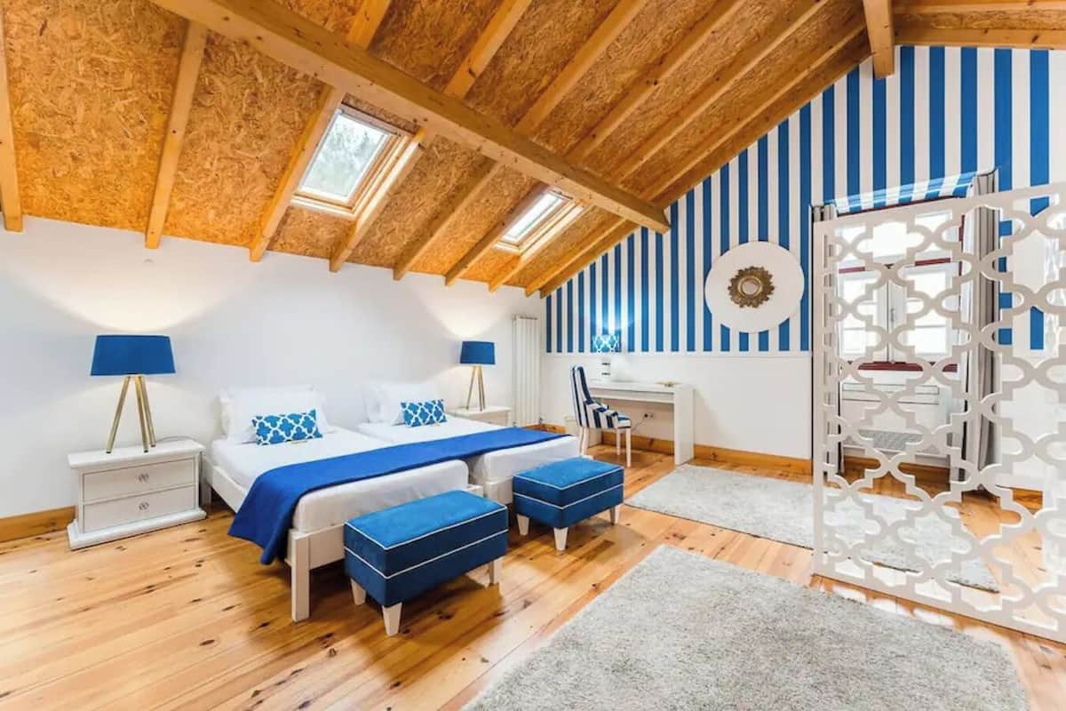 A bedroom with a sloped ceiling, wooden beams, a bed with blue bedding, a desk, and a chair.