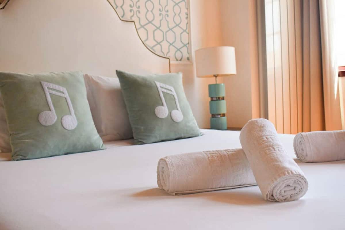 A hotel room with a bed, pillows with musical notes, a lamp, and rolled towels.