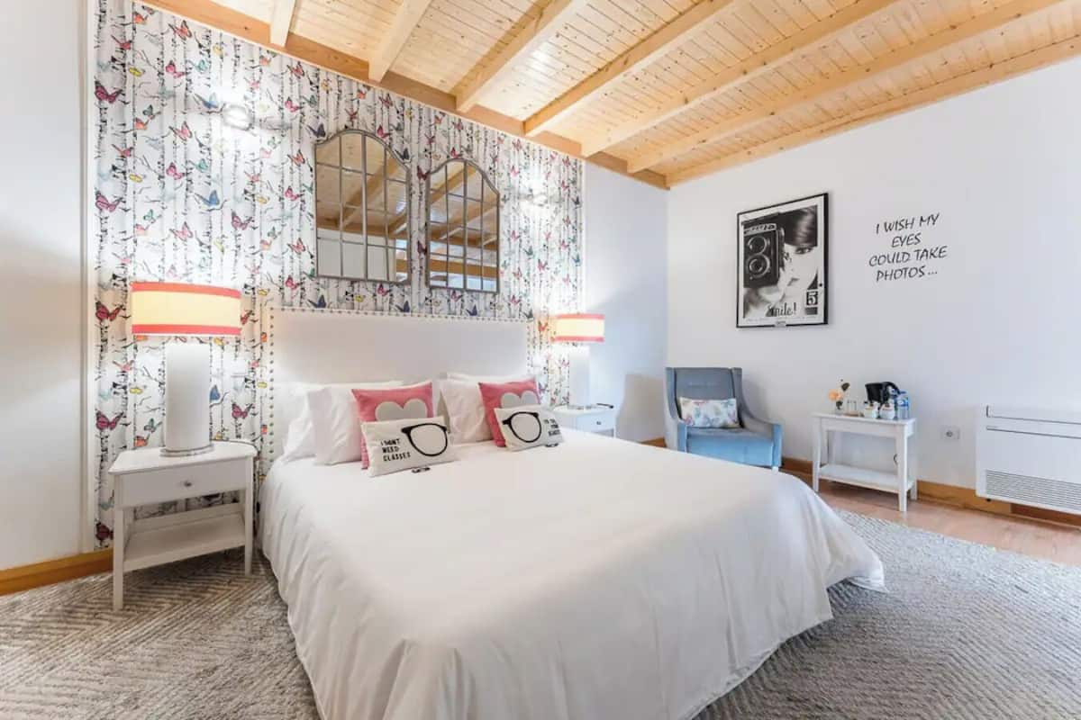 A bedroom with a large bed, two bedside tables, a chair, a mirror, and a wall with a quote.