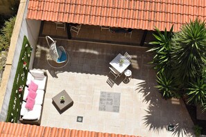 An outdoor patio with a tiled floor, a white sofa with pink cushions, a small table with a plant, and a round table with chairs.
