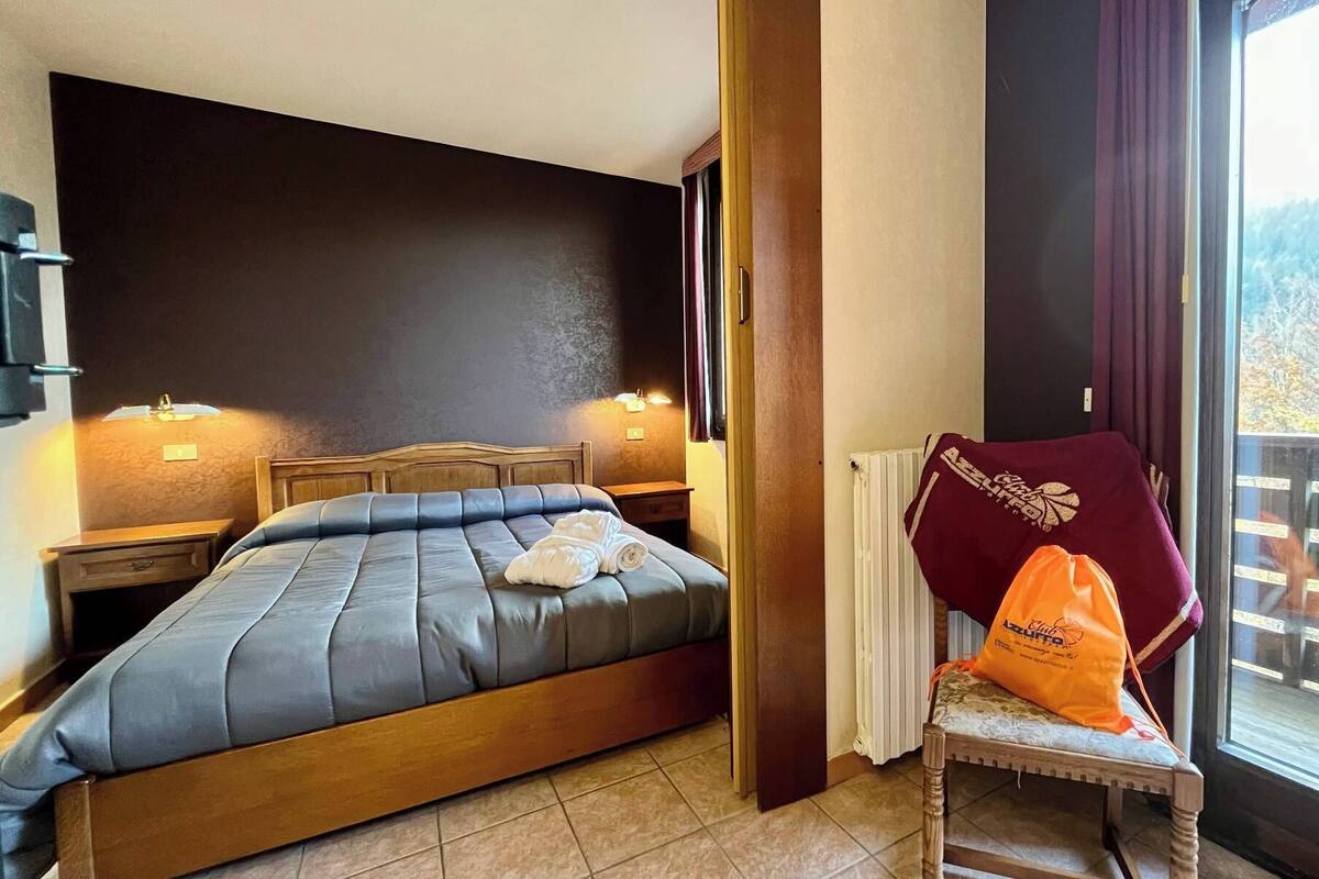 Une chambre d’hôtel avec un lit, deux lampes de chevet, une fenêtre donnant sur des arbres et une chaise avec un sac.