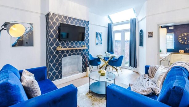 A modern living room with blue sofas, a glass coffee table, a fireplace with a geometric patterned wall, and a large window with curtains. - Spacious Bed Rest Stay in Salford City (Salford)