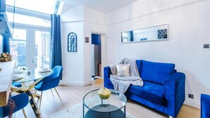 A modern living room with a blue sofa, a glass coffee table, and a dining area with blue chairs and a white table. - Spacious Bed Rest Stay in Salford City (Salford)
