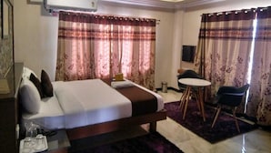 A hotel room with a bed, a desk, a chair, a small table, and a ceiling fan. - Green Valley Suites (Bengaluru)