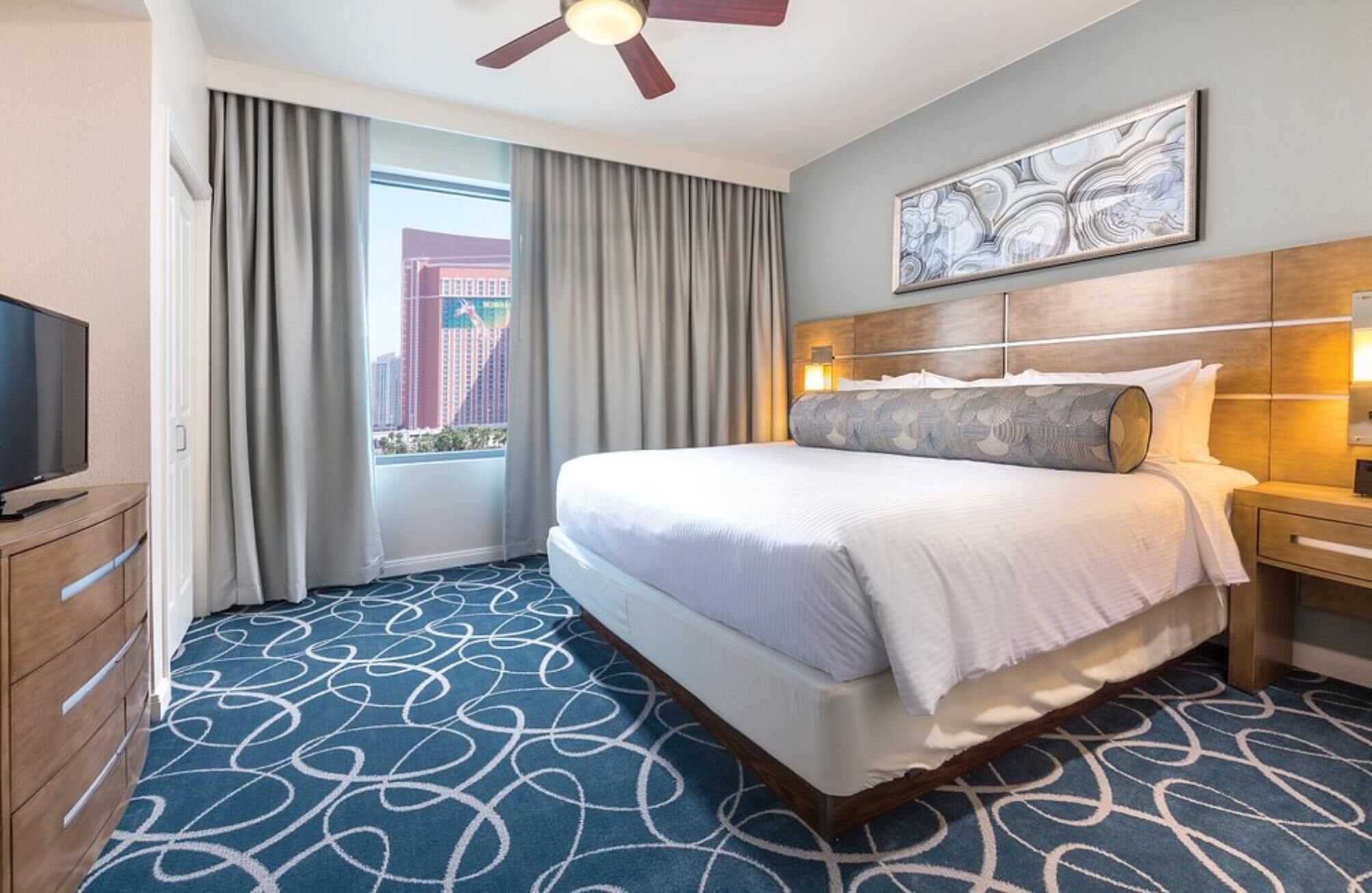 Een hotelkamer met een groot bed, een televisie, een plafondventilator, een raam met gordijnen en uitzicht over de stad.