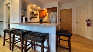 A modern kitchen with a central island, wooden stools, stainless steel appliances, and a 'HOME' sign on the wall.