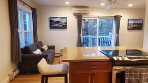 A compact living space with a kitchenette, a sofa, a chair, and a balcony with a view of the water.