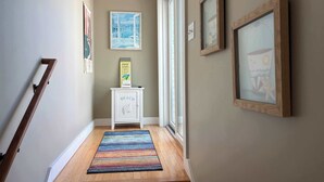 Un couloir doté d’un plancher en bois, d’un tapis rayé aux couleurs vives, d’une petite armoire blanche avec une inscription « Plage » et de tableaux encadrés accrochés aux murs.