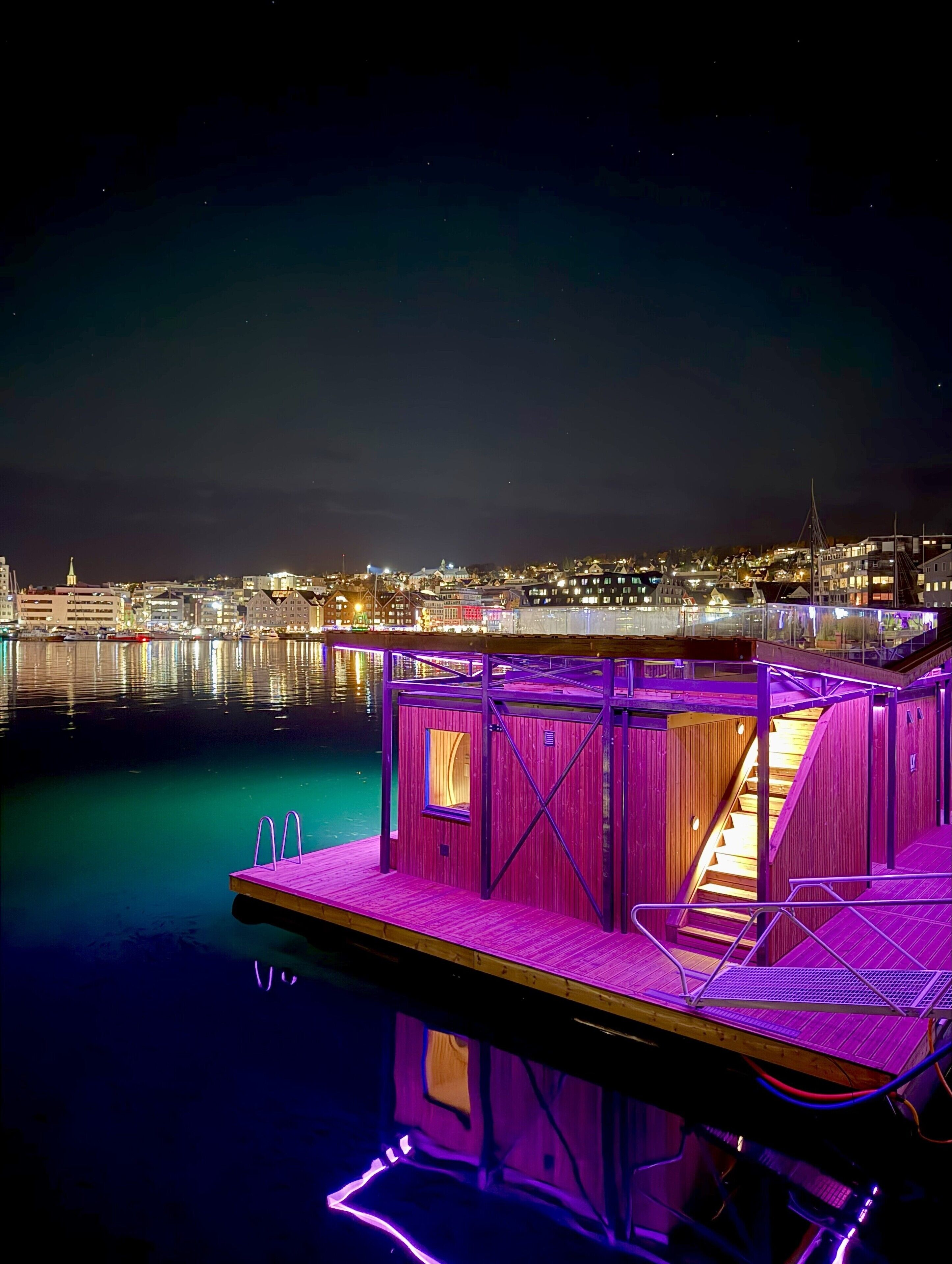 A docked boat with a purple-lit interior, a staircase, and a view of a cityscape at night.