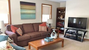 A living room with a brown sofa, a wooden coffee table, a television on a stand, and a bookshelf. - Ogunquit -2 Bedroom Family Condo with POOL 2 week stay (Ogunquit)