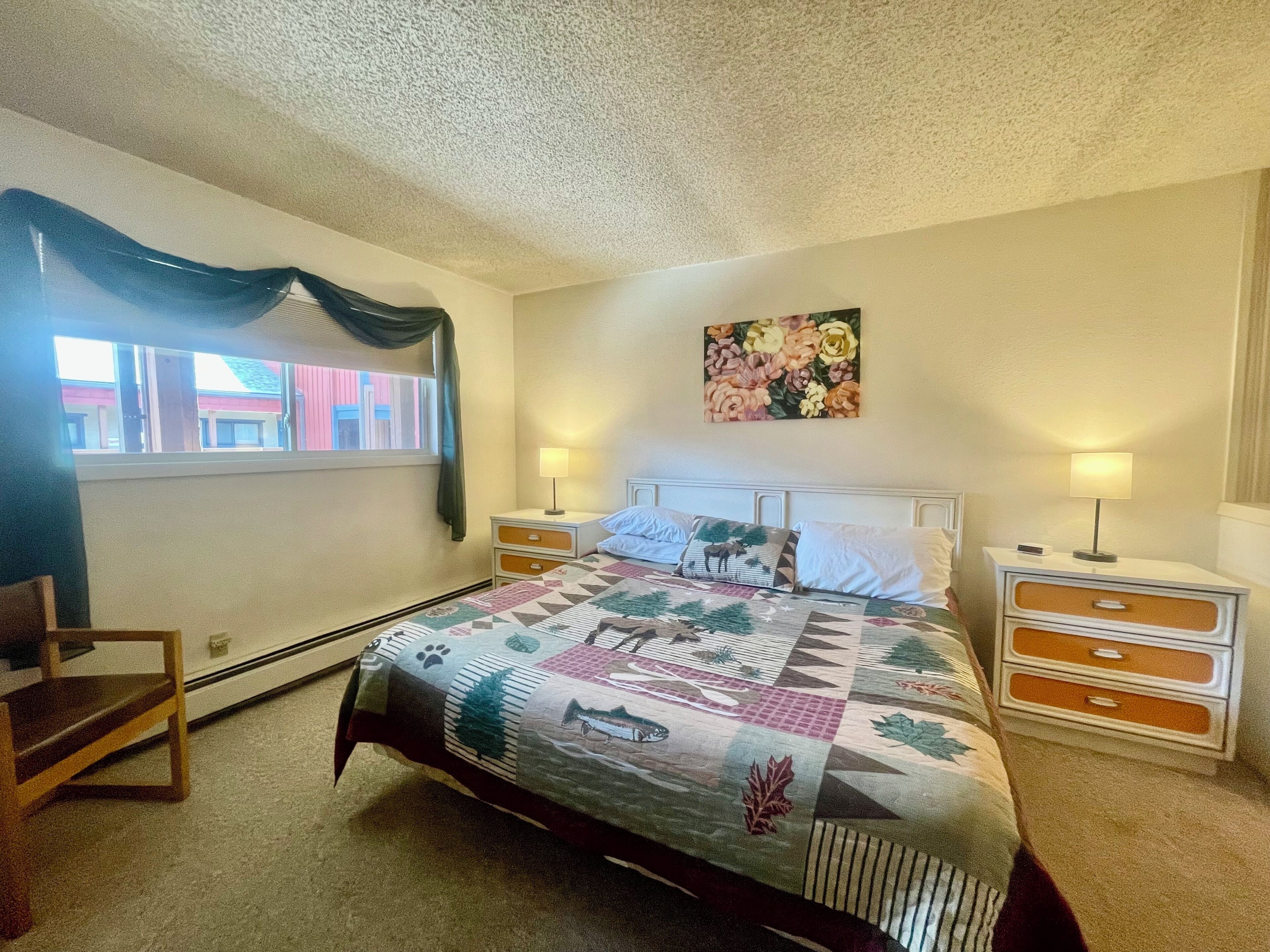 A bedroom with a bed, two bedside tables, a chair, a window with curtains, and a floral painting on the wall.