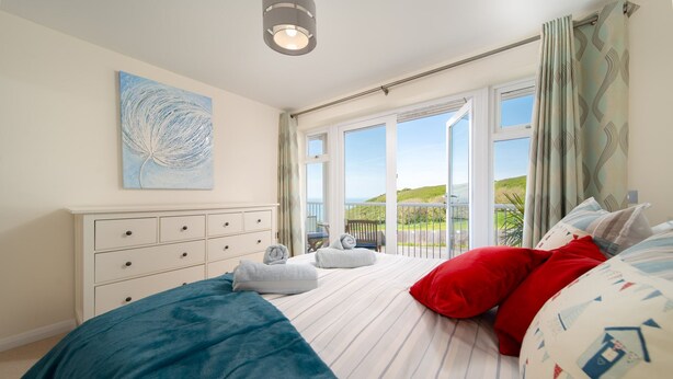 A bedroom with a bed, a dresser, a painting, and a balcony with a view. - Dog friendly, two bedroom holiday apartment with balcony in Mortehoe, North Devon. (Mortehoe)