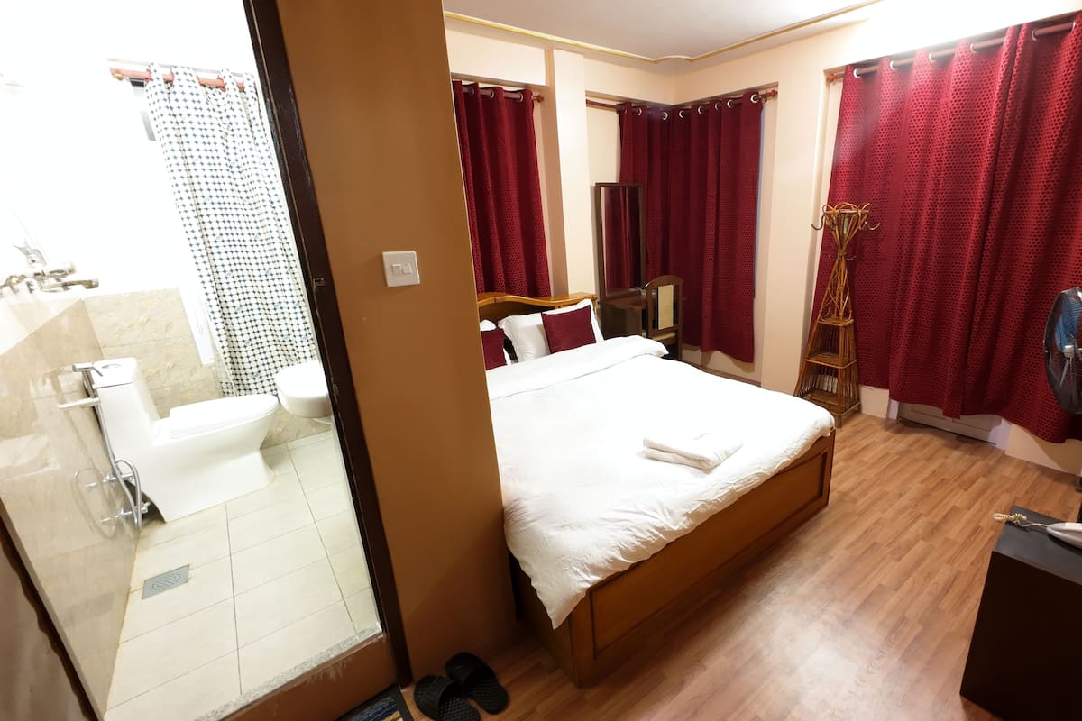 A hotel room with a bed, a bathroom with a shower, a fan, and a telephone.