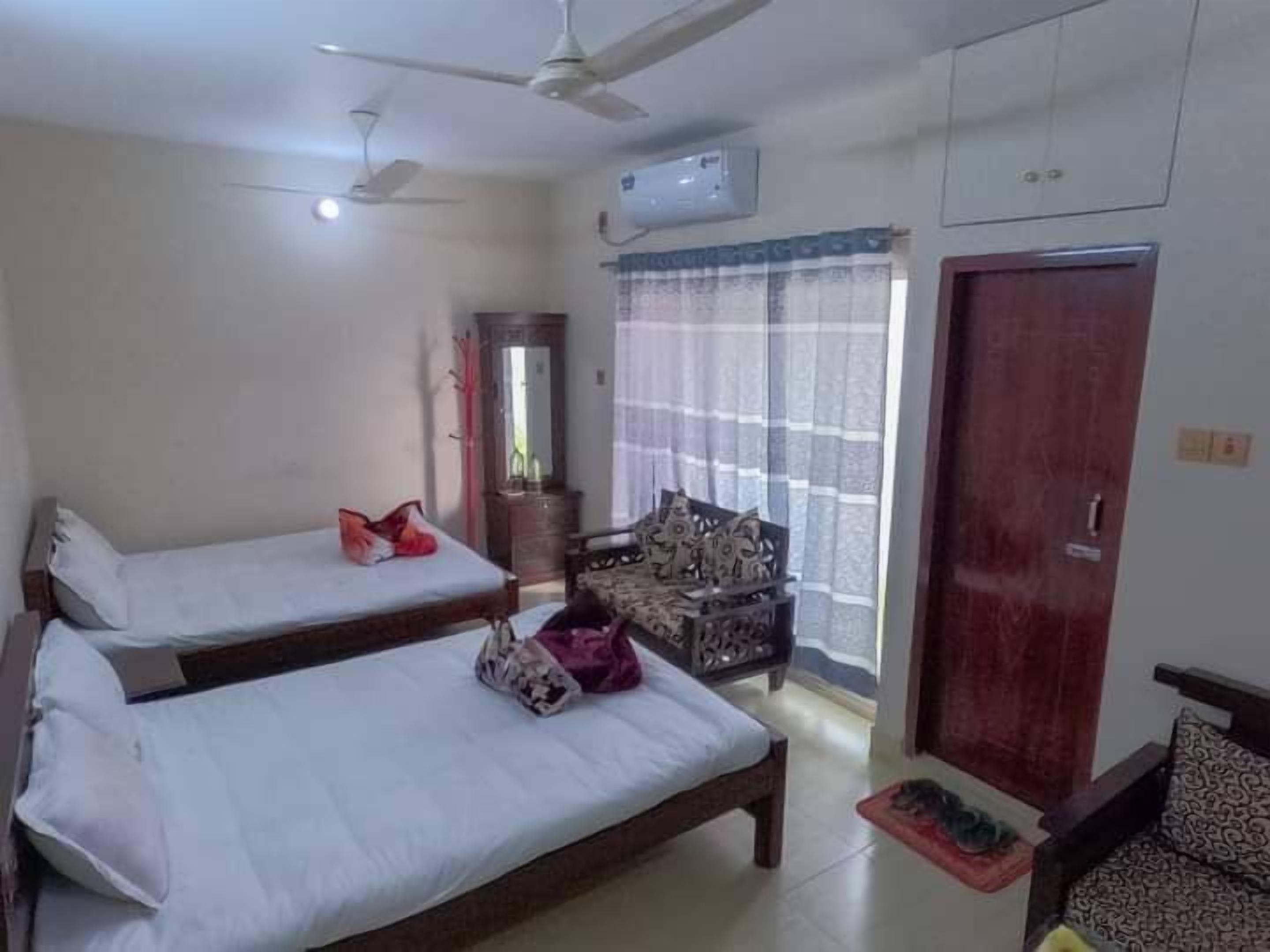 A hotel room with two single beds, a wooden wardrobe, a small table with a mat, a chair, a ceiling fan, and an air conditioning unit.