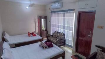A hotel room with two single beds, a wooden wardrobe, a small table with a mat, a chair, a ceiling fan, and an air conditioning unit.