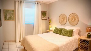 Un dormitorio con una cama grande, cortinas blancas, una mesita de noche con lámpara y dos adornos en la pared.