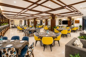A spacious dining area with multiple tables and chairs, a polished floor, and a high ceiling with wooden beams. - Hotel Le Premier (New Delhi)