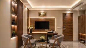 A modern living room with a flat-screen TV, two armchairs, a coffee table, and a wooden cabinet with shelves. - Hotel Le Premier (New Delhi)