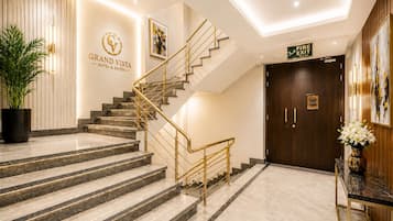 A hotel lobby with a marble staircase, a reception desk, a fire exit sign, and a large potted plant.