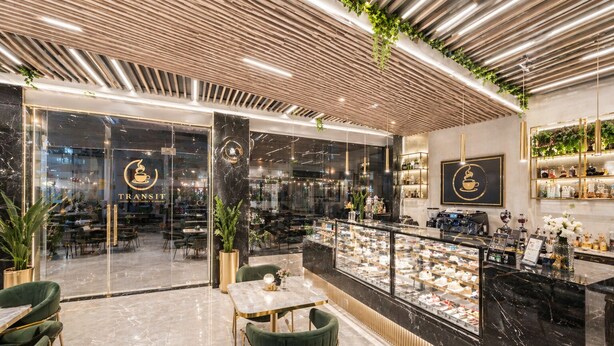 A modern coffee shop interior with marble flooring, green upholstered chairs, a glass display case, and a wooden ceiling. - Hotel Le Premier (New Delhi)