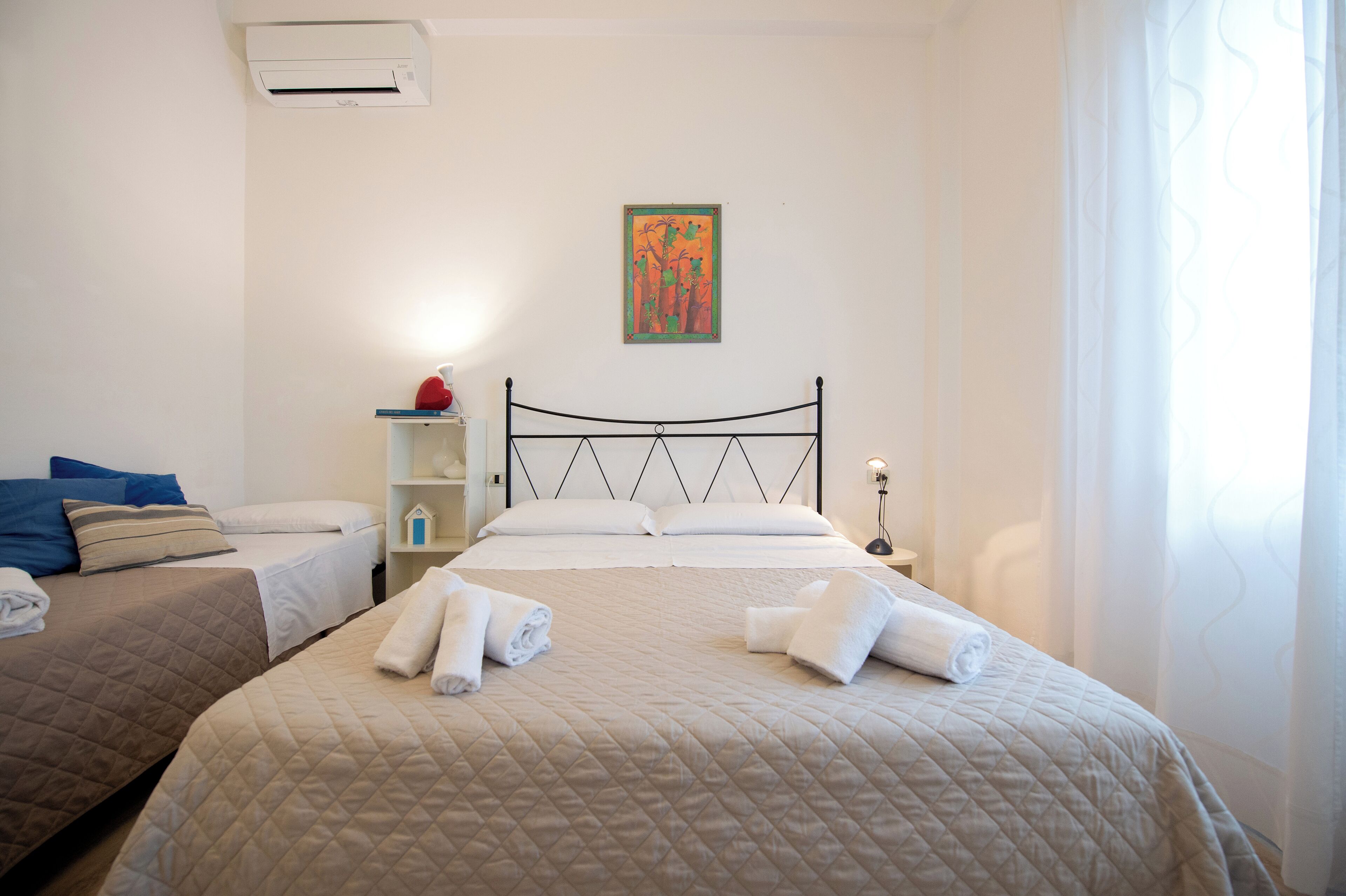 Hotellihuone, jossa on kaksi sänkyä, yöpöytä lampun kanssa, seinälle asennettu ilmastointilaite ja taulu.