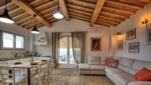 A spacious living area with a large beige sofa, a dining table set for four, and a kitchen with a white countertop and hanging pendant lights.