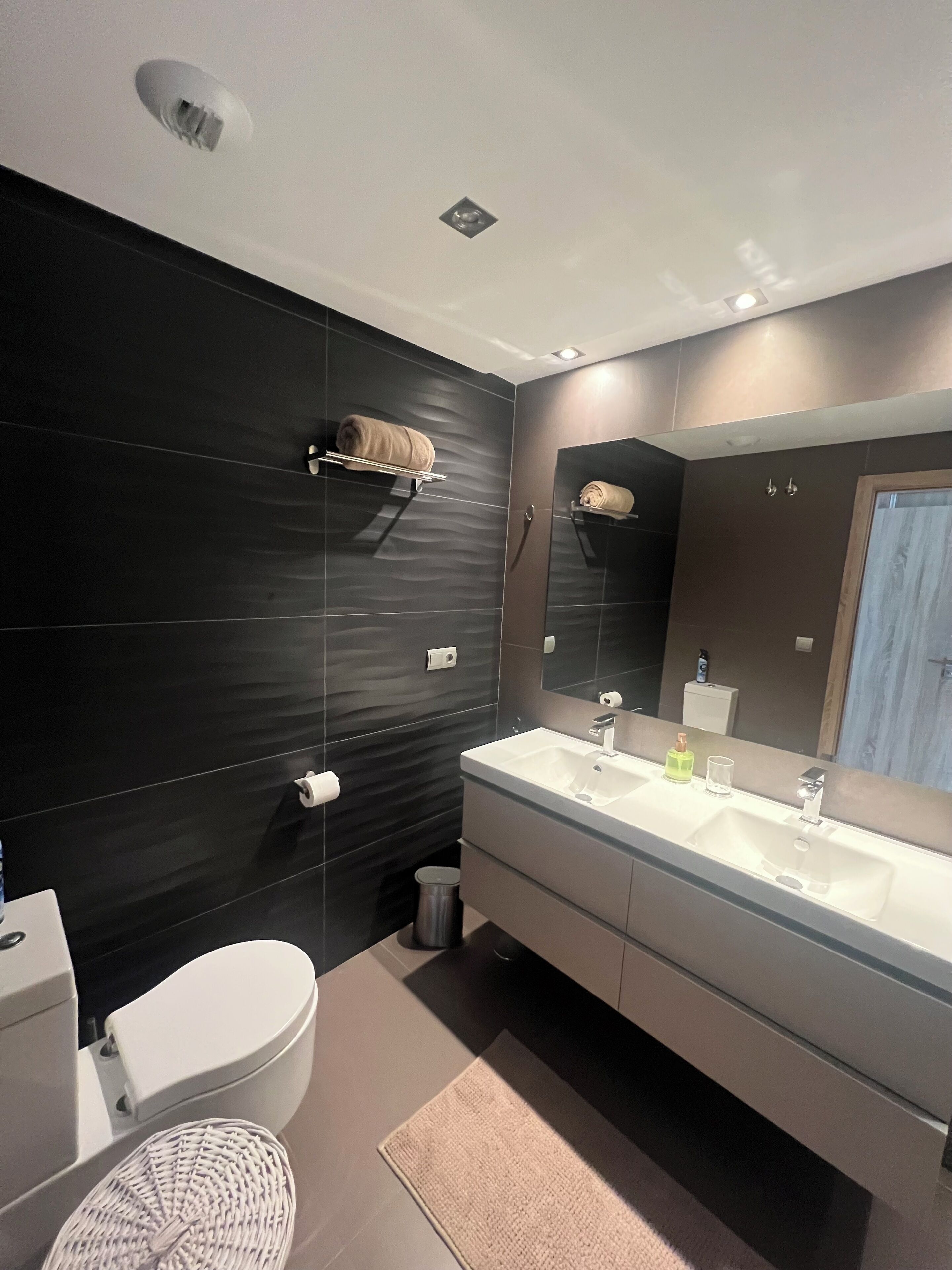 A modern bathroom with a dark tiled wall, a white sink with a mirror, a toilet, and a small trash bin.