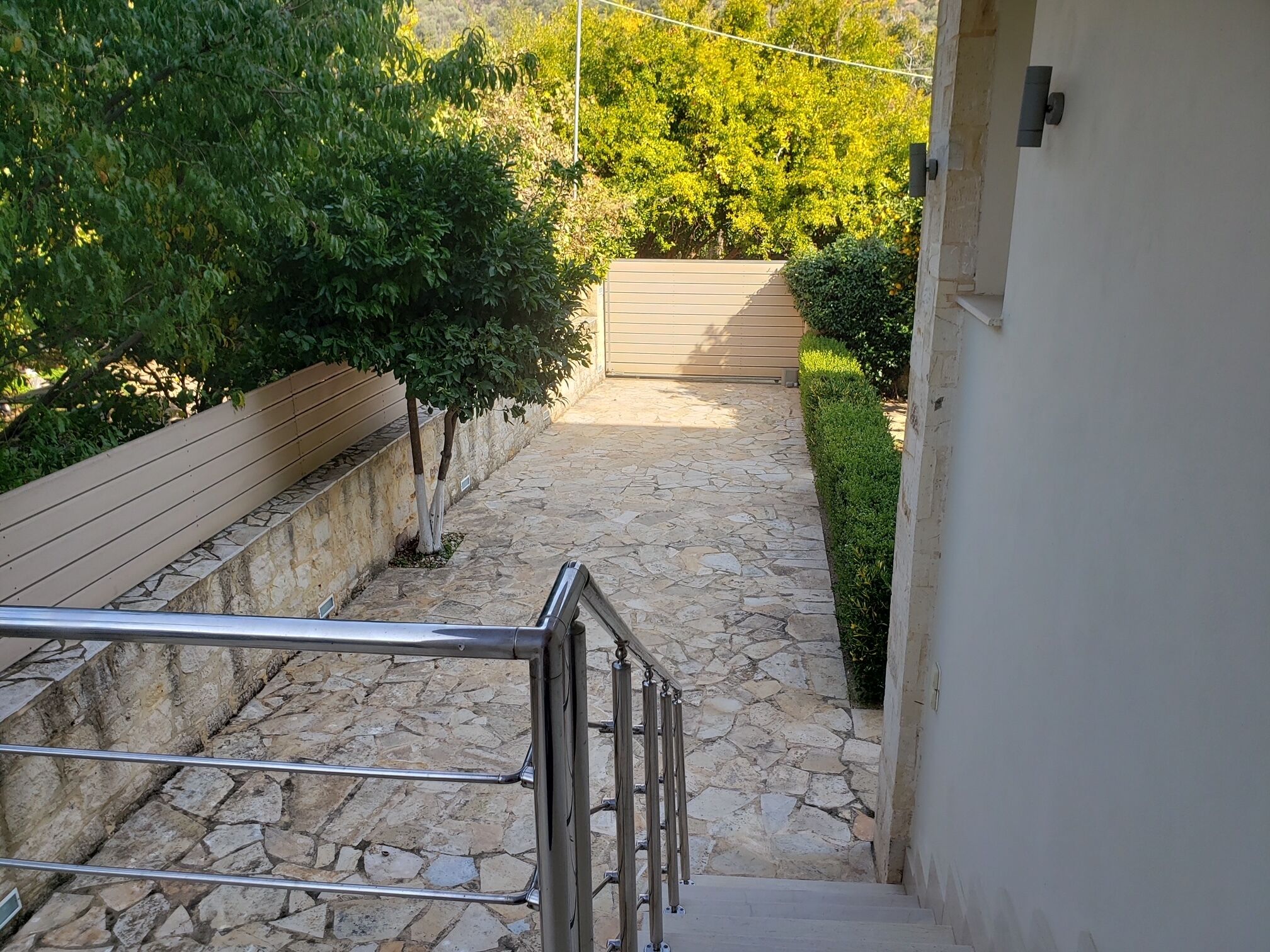 Un chemin de pierres menant à une porte en bois, bordé d’une rambarde en métal et d’un mur dont la base est en pierre.