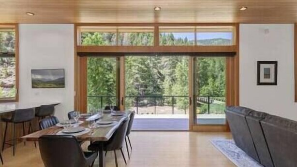 A dining area with a long table set for four, a sofa, and a view of trees through large windows. - Rio Vista Telluride w/ hot tub! (San Miguel County)