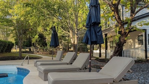 Zona junto a la piscina con sillones de descanso y sombrillas, rodeada de árboles y un edificio al fondo.