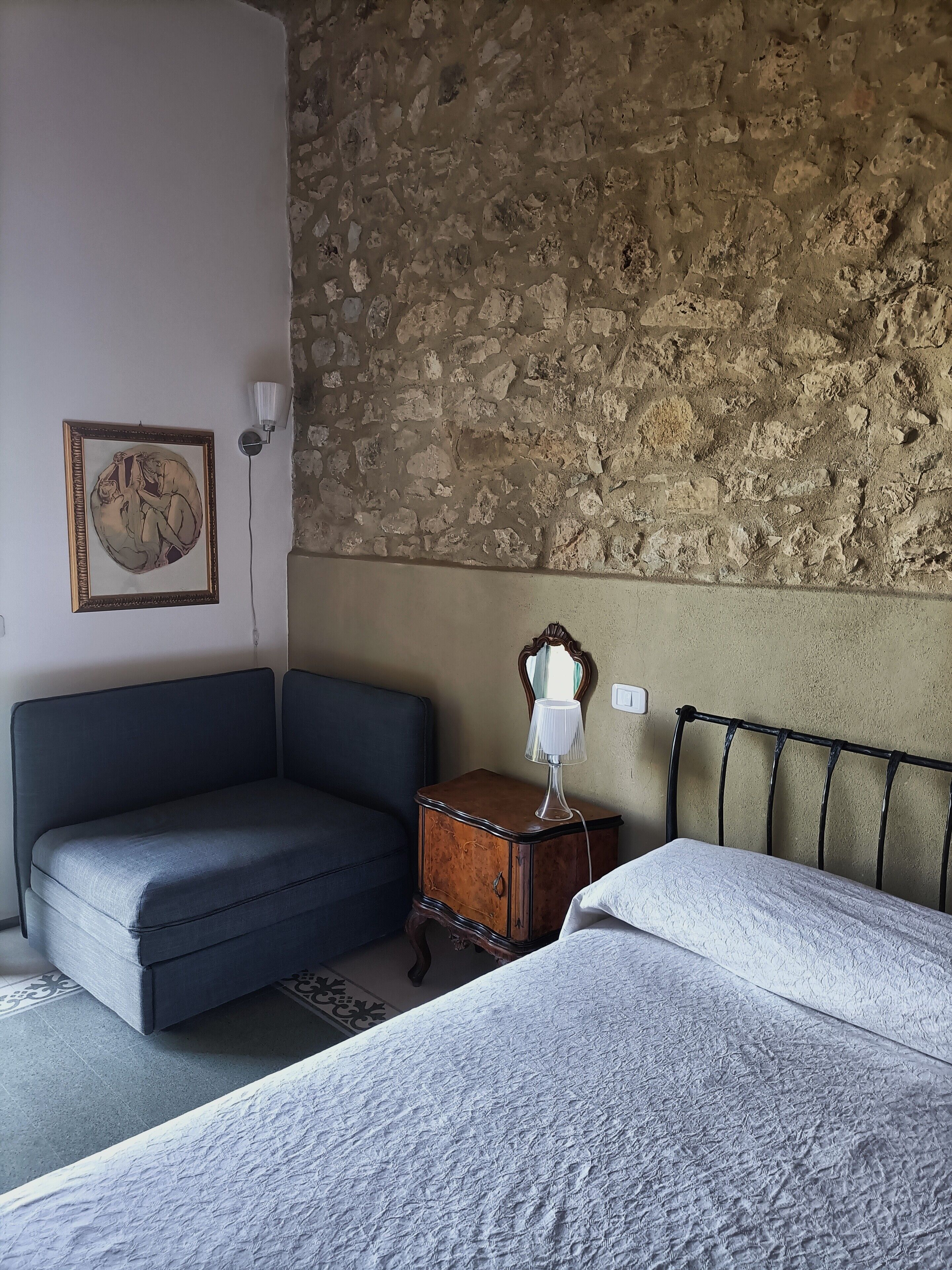 A room with a stone wall, a bed, a small table with a lamp, and a sofa.