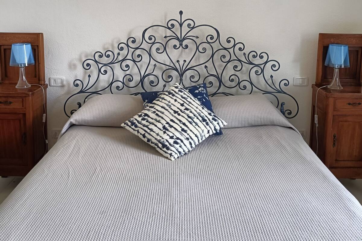 A neatly made bed with a striped pillow and two matching pillows, a wooden nightstand with a lamp, and a wall-mounted air conditioner.
