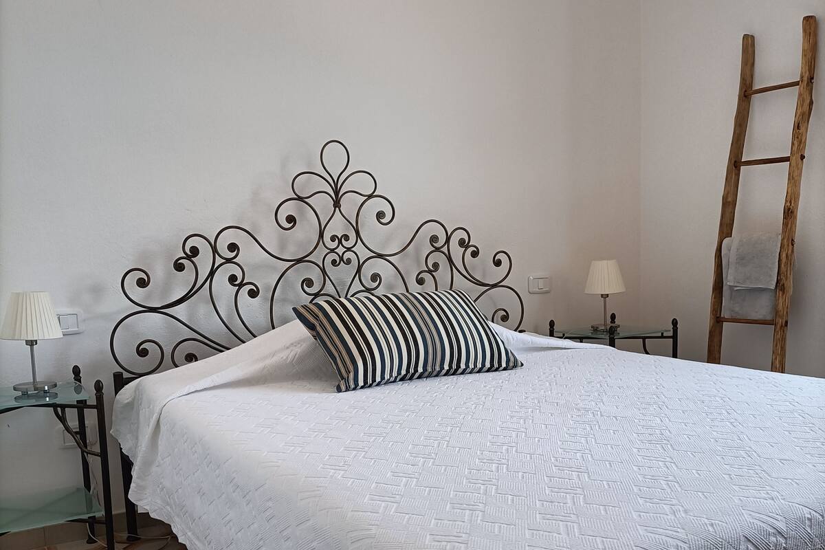 A neatly made bed with a white bedspread and a striped pillow, a metal headboard, a wooden ladder against the wall, and a patterned floor.