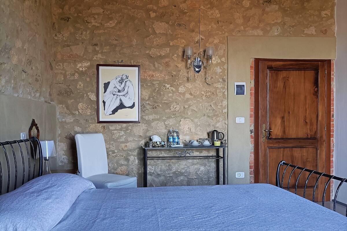 A bedroom with a large bed, a chair, a table with items, a wooden door, and a stone wall.