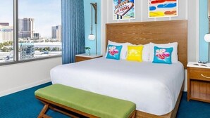 Habitación de hotel con una cama grande, un banco, una mesita de noche, una lámpara y vistas a la ciudad.