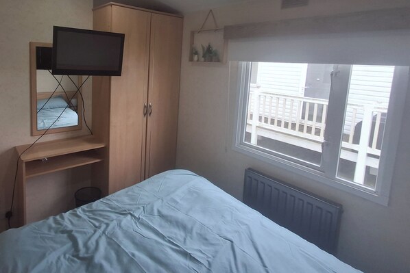 A bedroom with a bed, a TV mounted on a wooden cabinet, a window with a view of a balcony, and a radiator.