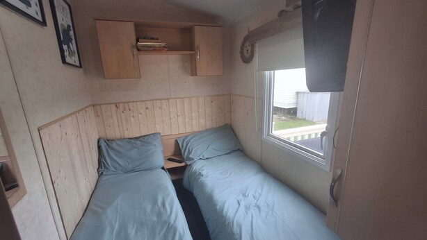 A compact bedroom with two single beds, a small shelf with books, and a window with a view of the outside. - 3-bed Caravan, Sleeps 8, Pool & Beach (Skegness)
