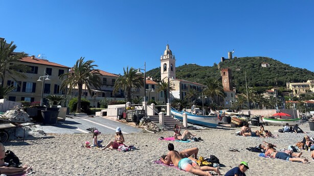 Une plage où des gens se prélassent au soleil, une église et une colline surmontée d’une tour.