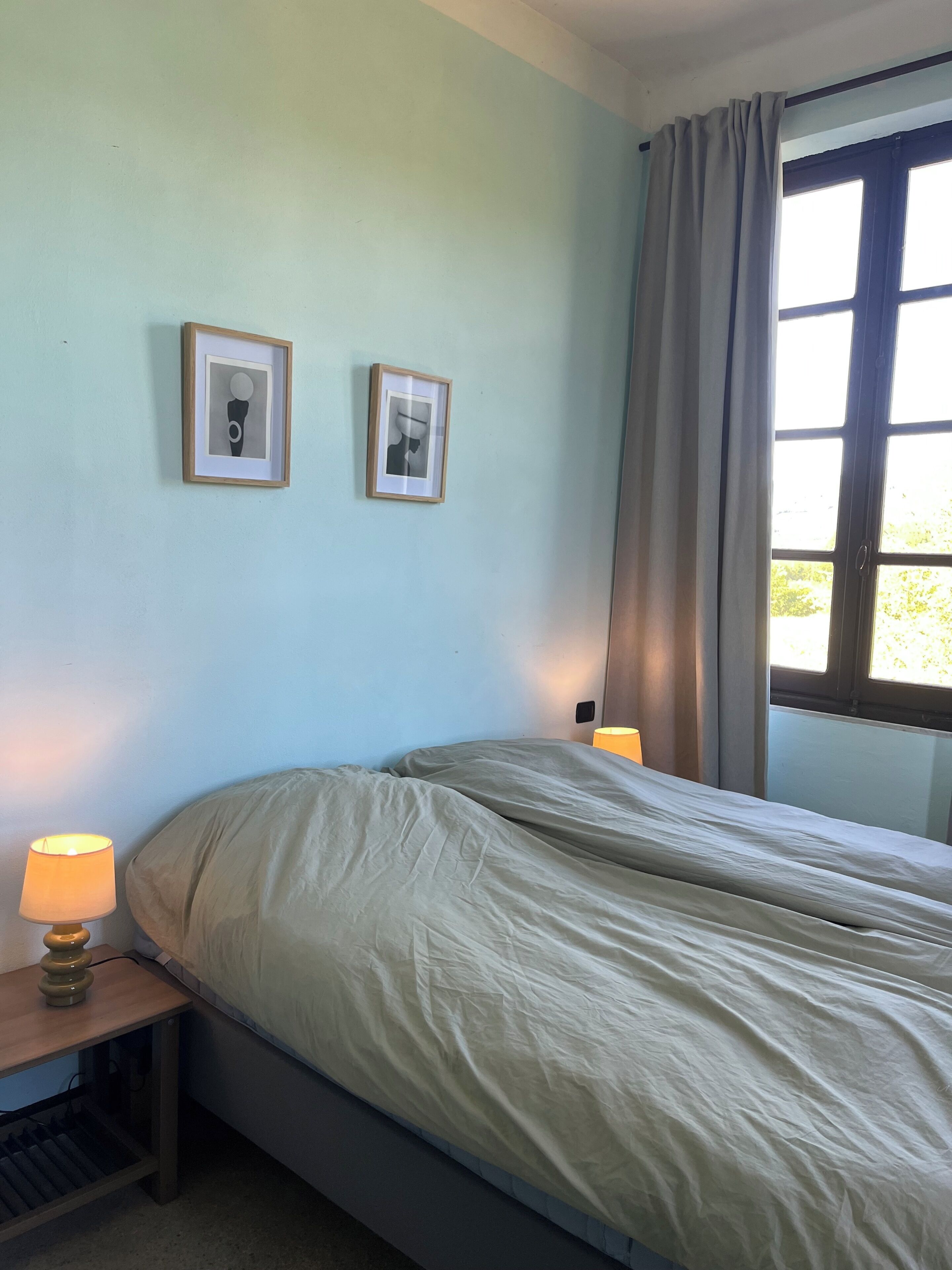 A bedroom with a bed, two framed pictures on the wall, a window with curtains, and a bedside lamp.