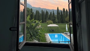 Une fenêtre ouverte donnant sur une piscine, un espace salon abrité et un paysage boisé.