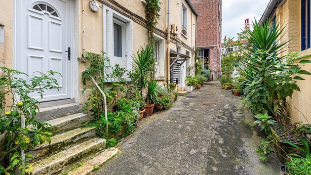 A narrow alleyway with steps leading to a white door, flanked by potted plants and a brick building in the background. - GuestReady - loft with workspace and music (Paris 19 Buttes-Chaumont)