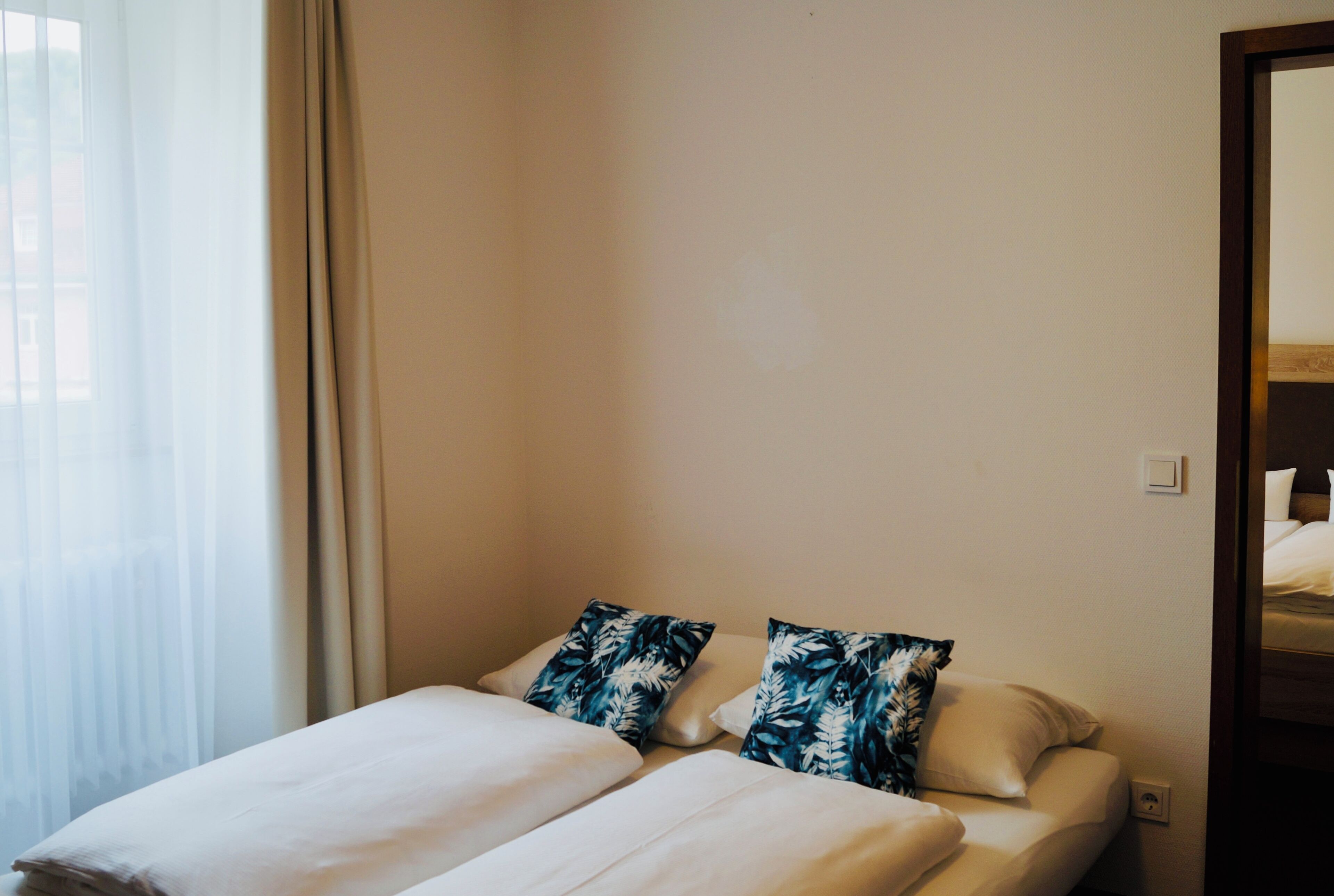 Ein Hotelzimmer mit einem Bett, zwei Kissen, einem Fenster mit Vorhängen und einer Tür.
