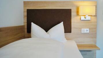A neatly made bed with white linens and a wooden headboard. A wall-mounted light fixture is visible on the right side of the headboard.