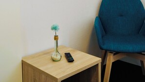 A room with a TV mounted on the wall, a small wooden table with a vase and remote control, a blue chair, and a wall clock. - Hotel & Restaurant Pelikan (Beuron)