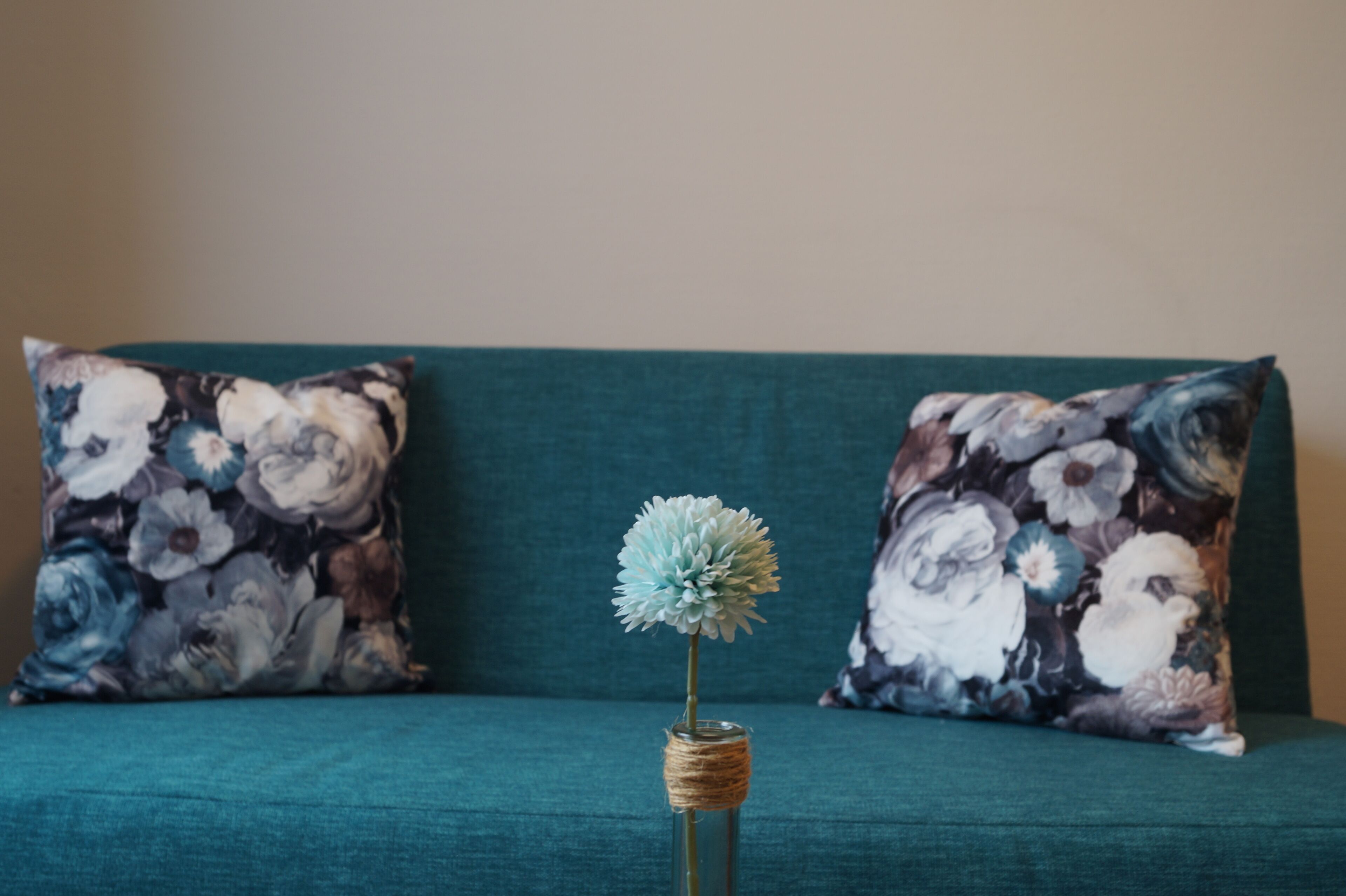 A teal sofa with floral-patterned cushions and a glass vase with a single flower.