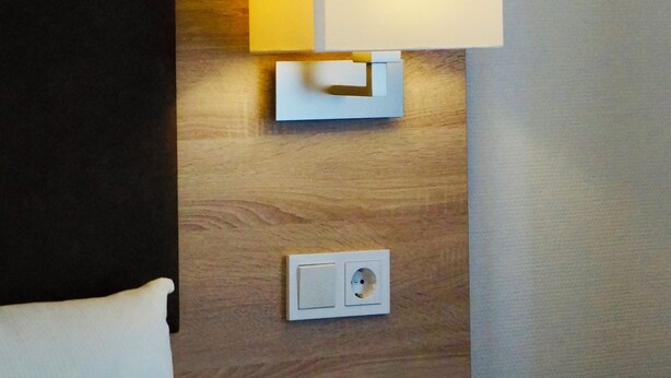 A hotel room with a wooden headboard, a white bed, a bedside table with a lamp, and a power outlet. - Hotel & Restaurant Pelikan (Beuron)